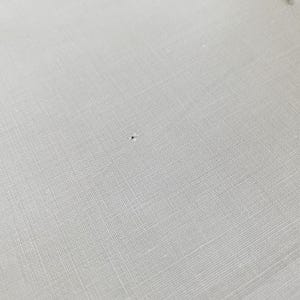 May include: A light gray, woven fabric textile featuring a visible, small, dark-colored puncture hole near the center. The material displays a consistent, fine-threaded grid pattern throughout the surface, highlighting the texture of the weave.
