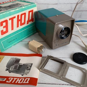 May include: Vintage slide projector with a teal and gray body, a lens, and a power cord. The box and manual are also visible, with Cyrillic text. A light bulb and slide holder are also included.