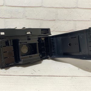 May include: A black vintage camera with its back open, displaying the internal mechanisms. The camera sits on a white wooden surface, with a brick wall backdrop. The interior of the camera is visible, including the film compartment.