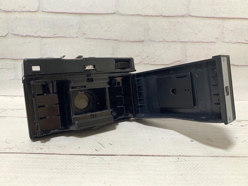 May include: A black vintage camera with its back open, displaying the internal mechanisms. The camera sits on a white wooden surface, with a brick wall backdrop. The interior of the camera is visible, including the film compartment.