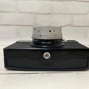 May include: A black vintage camera with a silver lens. The lens has numbers and markings, including "MADE IN USSR". The camera body is rectangular with a textured surface. The camera is on a white wooden surface.