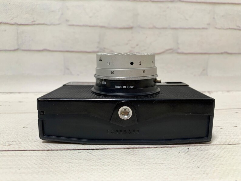 May include: A black vintage camera with a silver lens. The lens has numbers and markings, including "MADE IN USSR". The camera body is rectangular with a textured surface. The camera is on a white wooden surface.