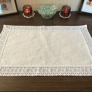 May include: A rectangular off-white placemat featuring a textured fabric center and a wide, intricate white crocheted border with a repeating circular pattern. The edges are finished with a delicate scalloped trim, creating a decorative frame.