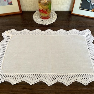 May include: A rectangular off-white linen placemat featuring a wide, intricate white crocheted lace border with a scalloped, zigzag edge. The center of the placemat is made of a smooth, plain-woven fabric, providing a contrast to the detailed lace.
