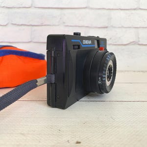 May include: A black vintage camera with the word "CMEHA" in blue lettering. The camera has a black strap and a red button. An orange and blue bag is in the background.