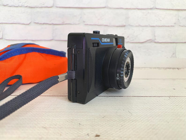 May include: A black vintage camera with the word "CMEHA" in blue lettering. The camera has a black strap and a red button. An orange and blue bag is in the background.