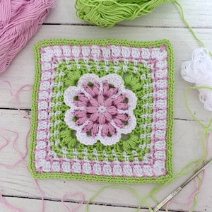 Könnte beinhalten: Ein gehäkeltes Quadrat mit einer rosa und weißen Blume in der Mitte. Das Quadrat ist von einem grünen Rand mit rosa und weißen Akzenten umgeben. Das Quadrat ist aus Garn und einer Häkelnadel gefertigt.