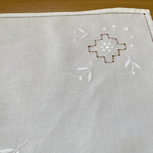 May include: A white rectangular linen napkin featuring a delicate embroidered geometric floral motif in the corner. The design includes white satin stitch petals and brown thread detailing, finished with a hemstitched border along the edges.