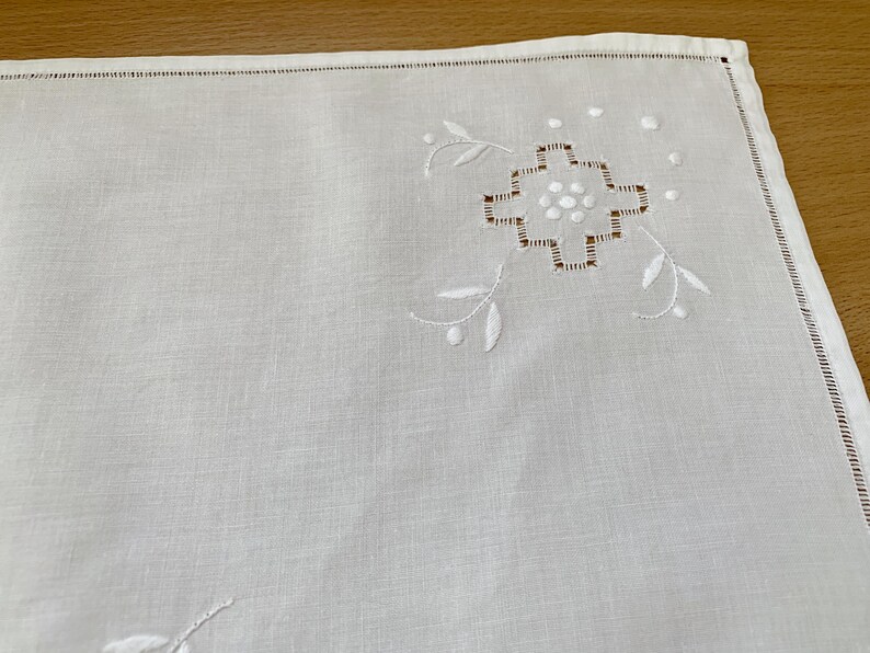May include: A white rectangular linen napkin featuring a delicate embroidered geometric floral motif in the corner. The design includes white satin stitch petals and brown thread detailing, finished with a hemstitched border along the edges.