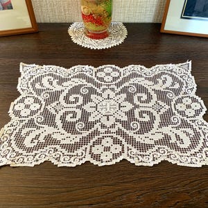 Può includere: Una tovaglietta rettangolare in pizzo bianco con un motivo floreale e a volute dettagliato. La tovaglietta è posizionata su una superficie di legno marrone scuro. I bordi sono smerlati, aggiungendo un tocco decorativo. È visibile anche un piccolo sottobicchiere rotondo in pizzo.