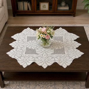 May include: A white square crochet doily featuring an intricate openwork pattern with geometric and floral motifs, centered on a dark brown wooden coffee table with a clear glass vase containing a bouquet of pink and white flowers.
