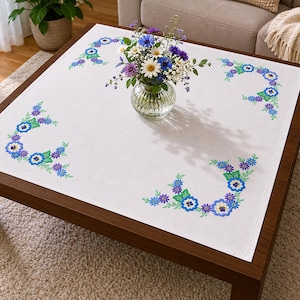 Puede incluir: Un mantel cuadrado de color blanco con patrones florales bordados en las esquinas con hilo azul, morado y verde. La tela tiene un borde dobladillado visible. Un florero de vidrio transparente con un ramo de flores blancas y azules se sitúa en el centro.