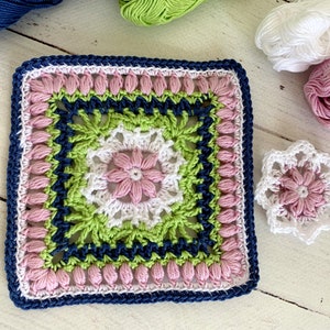 May include: A square, handmade crochet piece with a floral design in pink, white, green, and navy blue. A smaller, matching crochet flower is next to it. The yarn colors are visible in the background.