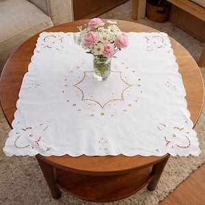 Può includere: Un tavolino rotondo in legno con una tovaglia bianca ricamata. Un vaso di vetro contiene un bouquet di fiori rosa e bianchi. La tovaglia ha un bordo smerlato e ritagli floreali, creando un aspetto delicato.