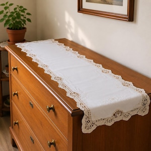 May include: A rectangular white table runner featuring a central fabric panel with subtle floral embroidery, bordered by an intricate beige crochet lace trim with scalloped edges, draped across the top surface of a wooden chest of drawers.