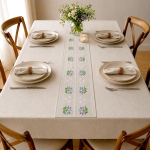 Könnte beinhalten: Eine beige rechteckige Tischdecke mit einem mittigen Läufer, der mit blauen und grünen Blumenmustern bestickt ist. Vier Gedecke sind mit weißen Tellern, in geflochtenen Ringen gefalteten Servietten und Silberbesteck angerichtet.