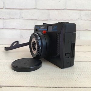 May include: A black vintage camera with the text "CIENA 35" in blue. The camera has a black lens cap and a black strap. The camera is on a light-coloured wooden surface.