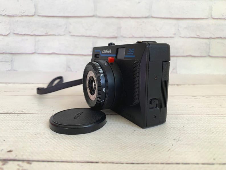 May include: A black vintage camera with the text "CIENA 35" in blue. The camera has a black lens cap and a black strap. The camera is on a light-coloured wooden surface.