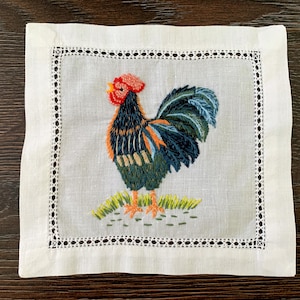 May include: Square white linen cocktail napkin with a detailed embroidered rooster design. The rooster features a red comb, orange and black body, and blue and green tail feathers. The napkin has a decorative, openwork border.