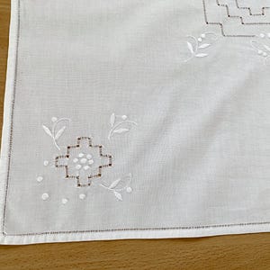 May include: A white rectangular textile featuring delicate white embroidery. The design includes geometric stepped patterns and small floral motifs with leaves and dots. The edges are finished with a straight hem and a decorative stitched border.