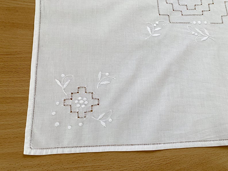 May include: A white rectangular textile featuring delicate white embroidery. The design includes geometric stepped patterns and small floral motifs with leaves and dots. The edges are finished with a straight hem and a decorative stitched border.