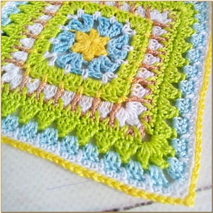 May include: A crocheted square with a colorful floral design in shades of green, yellow, blue, and white. The square has a border of yellow, white, and blue yarn.
