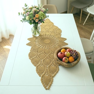 May include: A mustard yellow crochet table runner featuring an intricate, symmetrical leaf-like pattern that radiates from a central circular motif. The runner is laid flat on a white surface, accompanied by a glass vase of flowers and a wooden bowl.