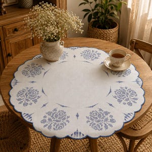 May include: A round white fabric tablecloth featuring a scalloped edge and a blue cross-stitch pattern of floral motifs arranged in a circular design. The tablecloth is placed on a wooden table with a ceramic vase of white flowers and a teacup.