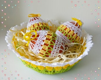 May include: Three crocheted Easter eggs in a white and green crocheted basket with yellow straw. The eggs are white with yellow and orange accents.