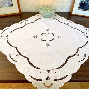 Könnte beinhalten: Ein weißes, quadratisches Deckchen mit einem komplizierten floralen und geometrischen Ausschnittmuster. Die Ränder sind gewellt, und die Mitte weist ein symmetrisches Blumenmuster auf. Das Deckchen liegt auf einer dunkelbraunen Holzoberfläche.