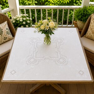 May include: A square white tablecloth featuring intricate grey geometric embroidery patterns and small floral motifs. The fabric is laid flat on a wooden table, centered with a clear glass vase containing a bouquet of white and yellow flowers.