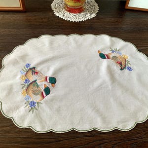 May include: White oval-shaped embroidered table runner with a scalloped edge. The runner features embroidered mallard ducks with green heads, brown bodies, and red accents, surrounded by blue and yellow floral details.