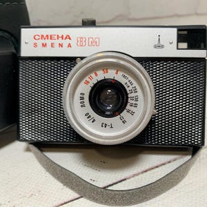 May include: A vintage black and silver Smena 8M camera with a textured body and a silver lens. The lens has various settings and markings. A grey strap is attached. A black case is visible on the left.