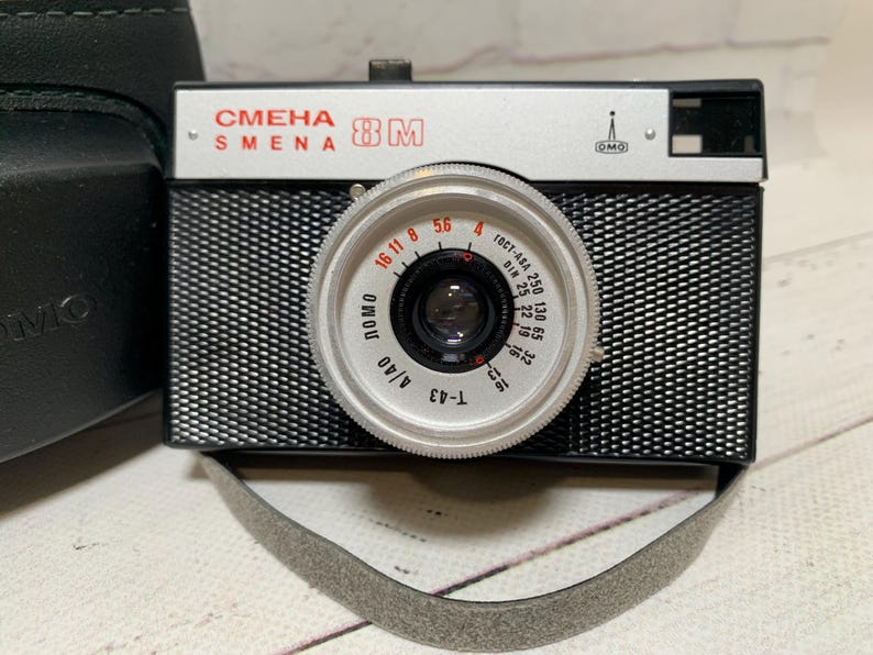 May include: A vintage black and silver Smena 8M camera with a textured body and a silver lens. The lens has various settings and markings. A grey strap is attached. A black case is visible on the left.
