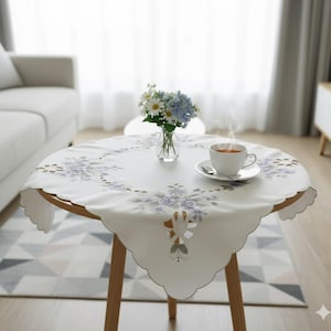 Puede incluir: Una mesa redonda de madera con un mantel blanco con un diseño floral en tonos morados. Una taza de té y un jarrón con flores están sobre la mesa. La mesa está en un salón.