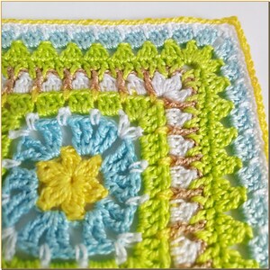 May include: Close-up of a crocheted square with a yellow, blue, and green floral design. The square is bordered with a white and yellow trim.