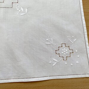 May include: A white rectangular textile featuring delicate white embroidery in the corners. The design includes a geometric cross-stitch pattern surrounded by small circular dots and stylized leaf motifs, finished with a simple hemmed edge.