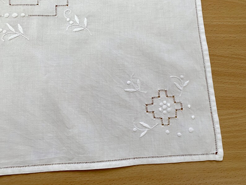 May include: A white rectangular textile featuring delicate white embroidery in the corners. The design includes a geometric cross-stitch pattern surrounded by small circular dots and stylized leaf motifs, finished with a simple hemmed edge.