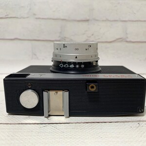 May include: A black vintage camera with a silver lens. The lens has various settings and markings. The camera body has a flash mount and a silver dial. The word "Smena" is printed in red on the top of the camera.