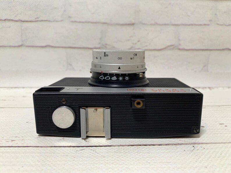 May include: A black vintage camera with a silver lens. The lens has various settings and markings. The camera body has a flash mount and a silver dial. The word "Smena" is printed in red on the top of the camera.