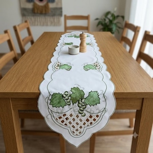 May include: A white table runner featuring scalloped edges and an embroidered pattern of green grape clusters and leaves. The design includes a lattice-style cutout detail at the ends. A wooden pepper mill and a small white bowl sit on the runner.