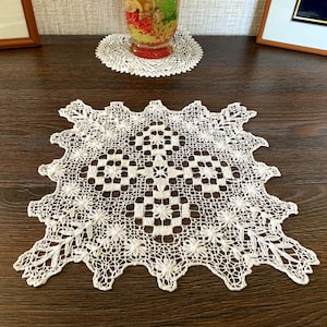 May include: A square off-white lace doily featuring an intricate needlework pattern with central geometric blocks, floral-inspired star motifs, and a scalloped border with delicate openwork detailing throughout the entire textile piece.