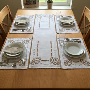 May include: A white linen table runner and four matching placemats featuring brown floral embroidery and scalloped edges. Each place setting includes a white ceramic plate, a folded beige napkin, a clear glass, a silver fork, and a silver spoon.