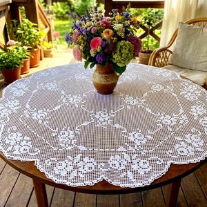 May include: A round white crochet tablecloth featuring an intricate floral pattern with scalloped edges. The tablecloth is draped over a circular wooden table, which holds a brown ceramic vase filled with a colorful bouquet of fresh flowers.