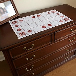 May include: A rectangular white table runner featuring a grid pattern of thin brown lines and embroidered dark red snowflake motifs arranged in a border along the edges. The fabric has a visible woven texture and is laid flat on a wooden surface.