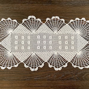 May include: A rectangular white crochet doily featuring a central grid pattern of small squares surrounded by a decorative border of repeating fan-shaped motifs with scalloped edges, crafted from fine thread with an intricate openwork design.