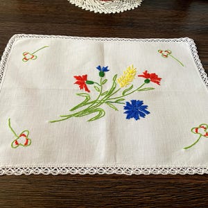 May include: A rectangular white fabric doily featuring a central embroidered bouquet of red, blue, and yellow wildflowers with green stems. The edges are finished with a white crocheted lace border, and four small clover motifs are embroidered in the corners.