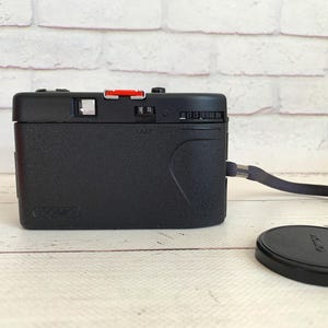 May include: A black Canon Sure Shot camera with a red button and a lens cap. The camera has a wrist strap attached and is on a light-coloured wooden surface.