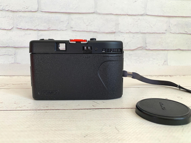 May include: A black Canon Sure Shot camera with a red button and a lens cap. The camera has a wrist strap attached and is on a light-coloured wooden surface.