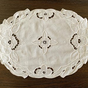 Puede incluir: Camino de mesa ovalado blanco con diseños calados de flores y hojas. El camino de mesa presenta grandes motivos florales en las esquinas y un diseño central en forma de estrella. Los bordes están festoneados, añadiendo un toque delicado.